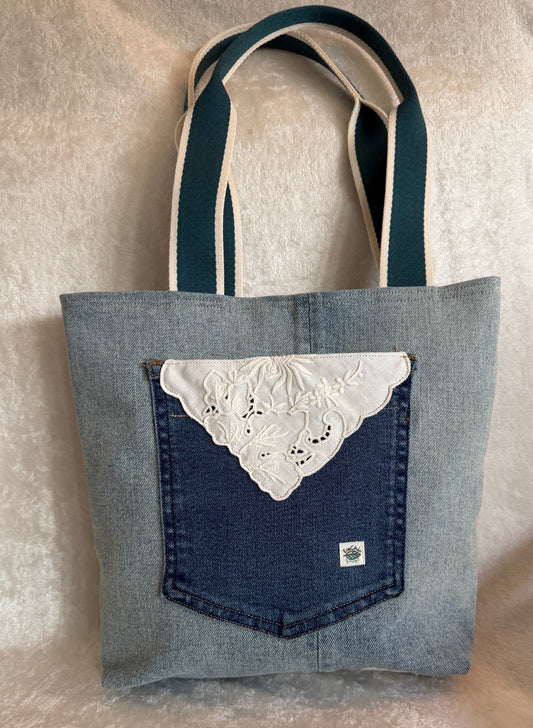 The back of a tote bag made from light blue upcycled denim fabric, featuring a darker blue jeans pocket with a white vintage linen cut-work applique.