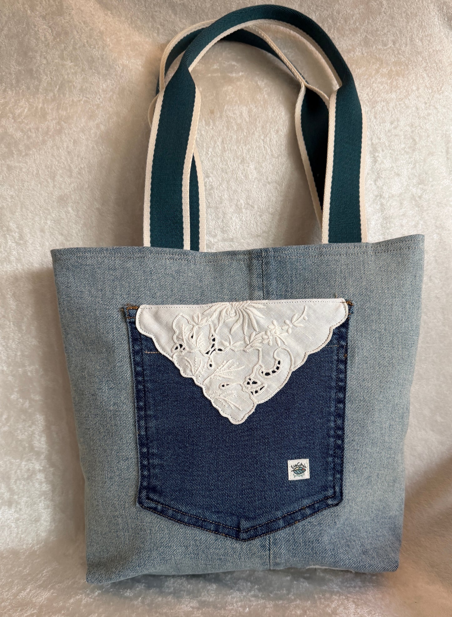 The back of a tote bag made from light blue upcycled denim fabric, featuring a darker blue jeans pocket with a white  vintage linen cut-work applique.