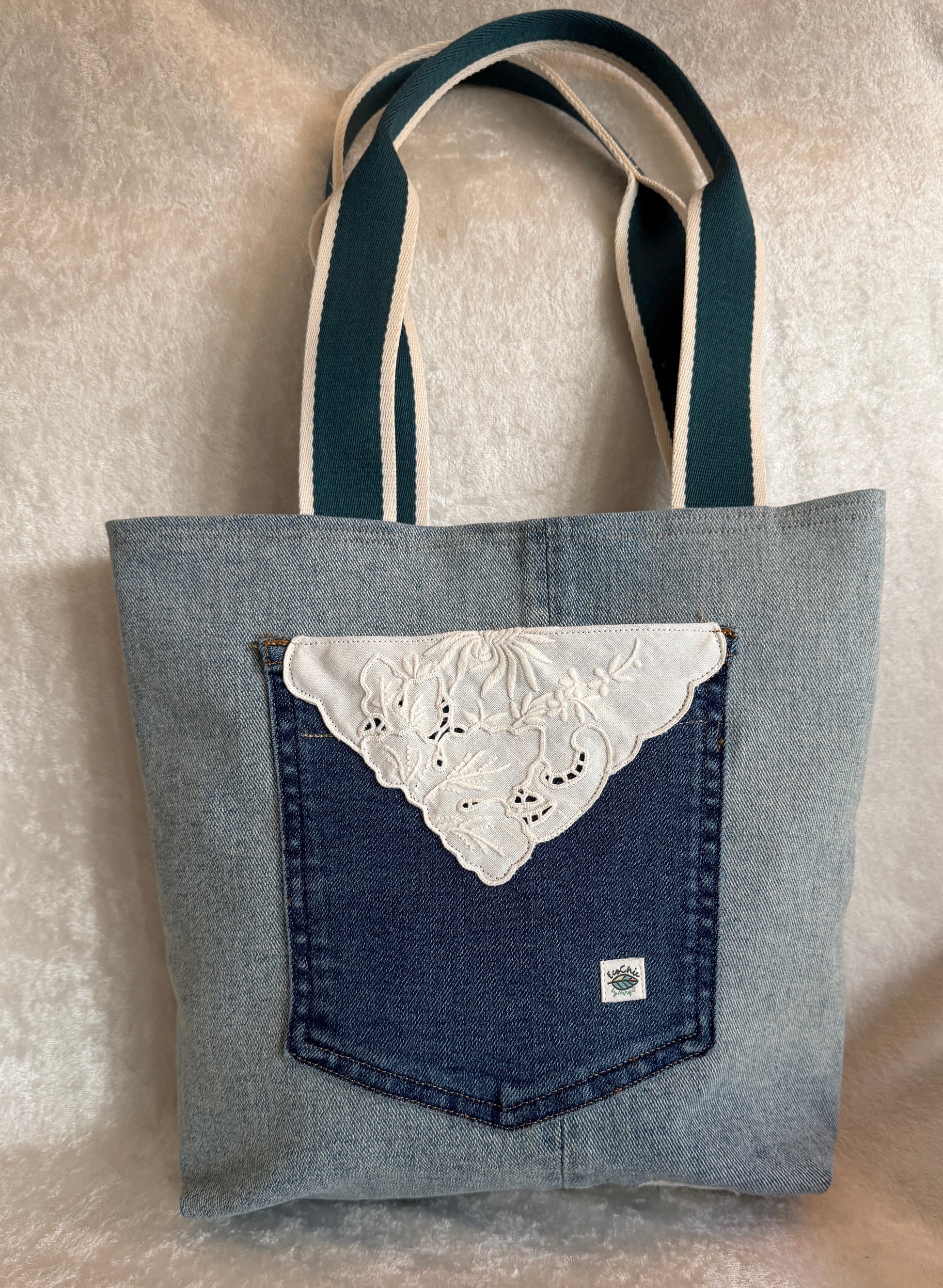 The back of a tote bag made from light blue upcycled denim fabric, featuring a darker blue jeans pocket with a white  vintage linen cut-work applique.