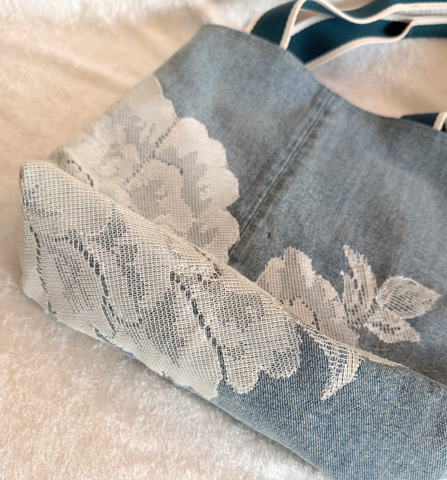 A close-up of the bottom of a light blue denim tote bag with floral lace applique. 