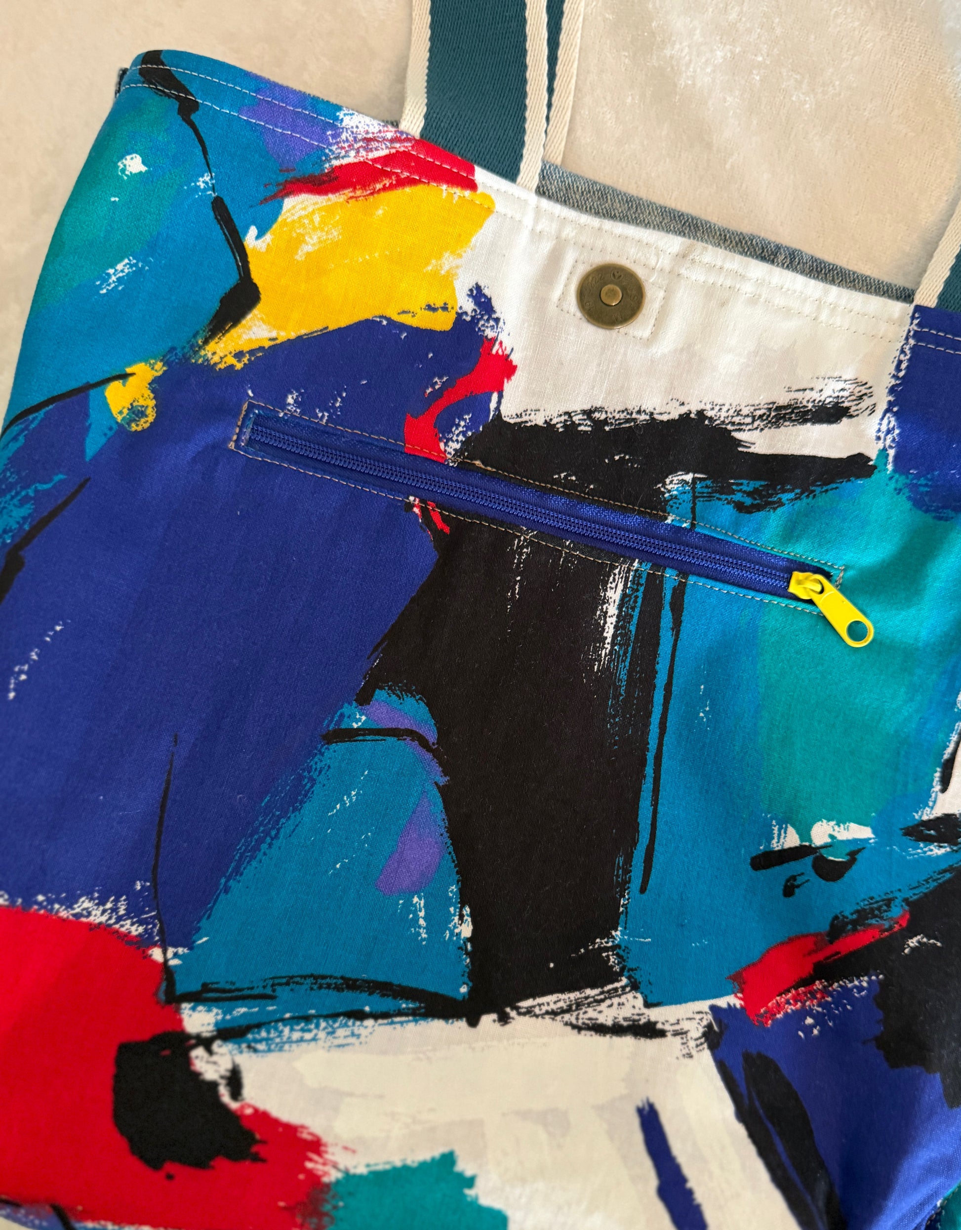 A close-up of the interior of an upcycled denim tote bag featuring. The cotton lining is an abstract, colorful brushstroke print. IT is easy to locate the  yellow zipper pull for the inside pocket.