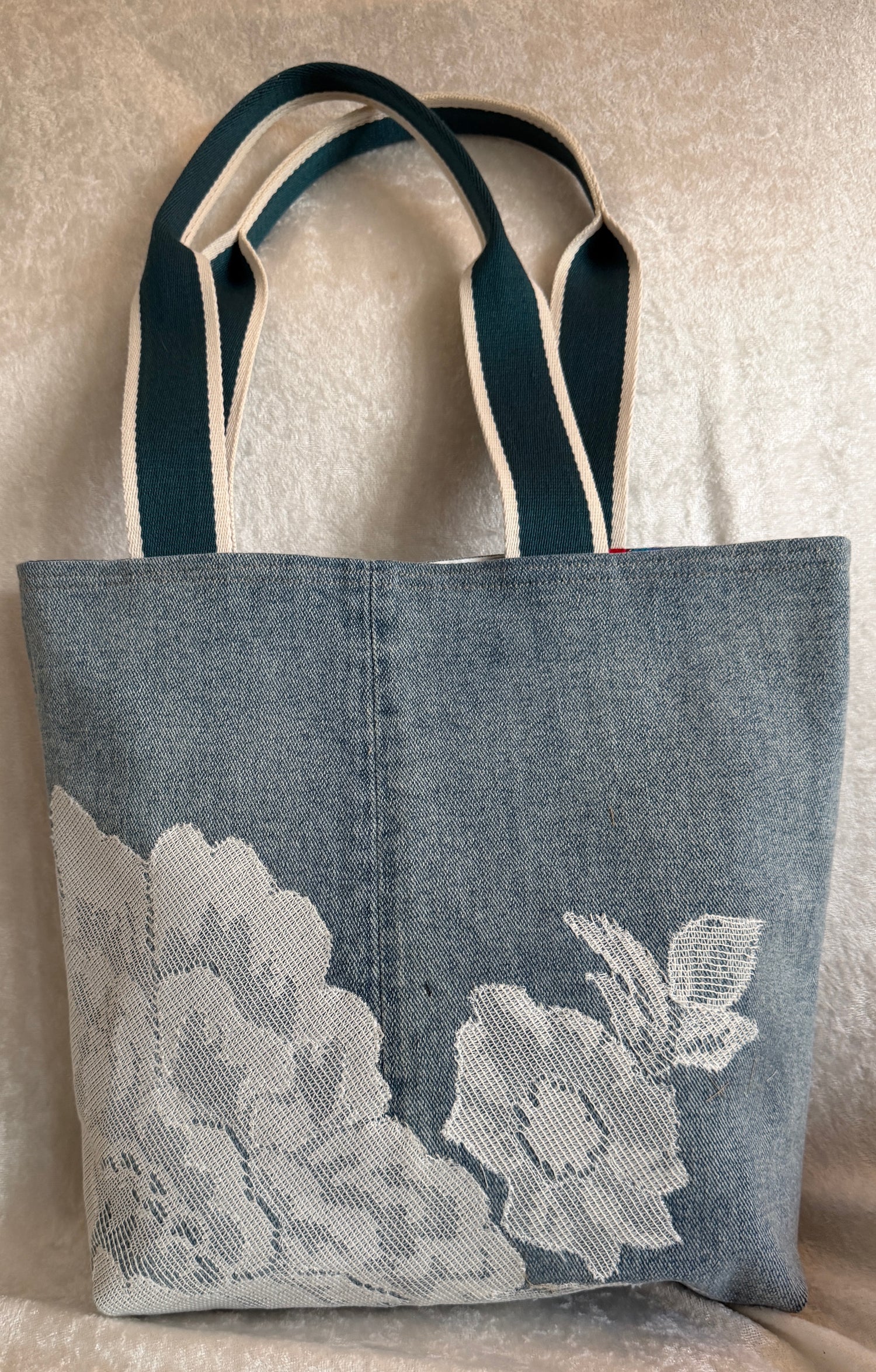A blue denim tote bag with white floral lace on the lower half and woven cotton shoulder straps. Denim and lace are upcycled.