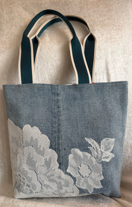 A blue denim tote bag with white floral lace on the lower half and woven cotton shoulder straps. Denim and lace are upcycled.